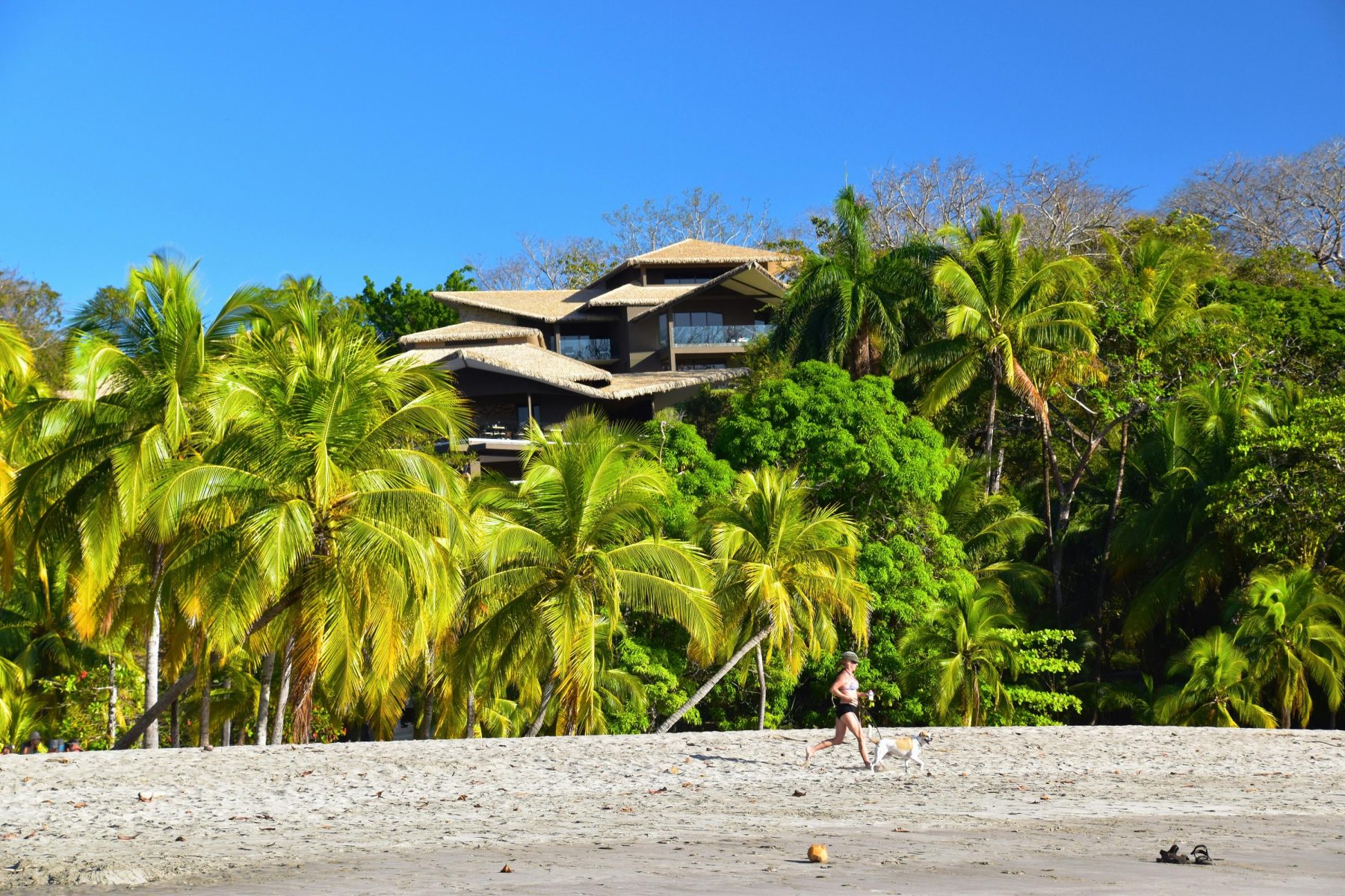 Een vrouw rennend met haar hond over het strand Playa Carrillo met op de achtergrond een groot huis, verscholen achter bomen. 