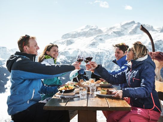 Vier mensen zitten aan een picknicktafel vol eten in een bergachtig, winters landschap, terwijl ze proosten met een glas wijn.