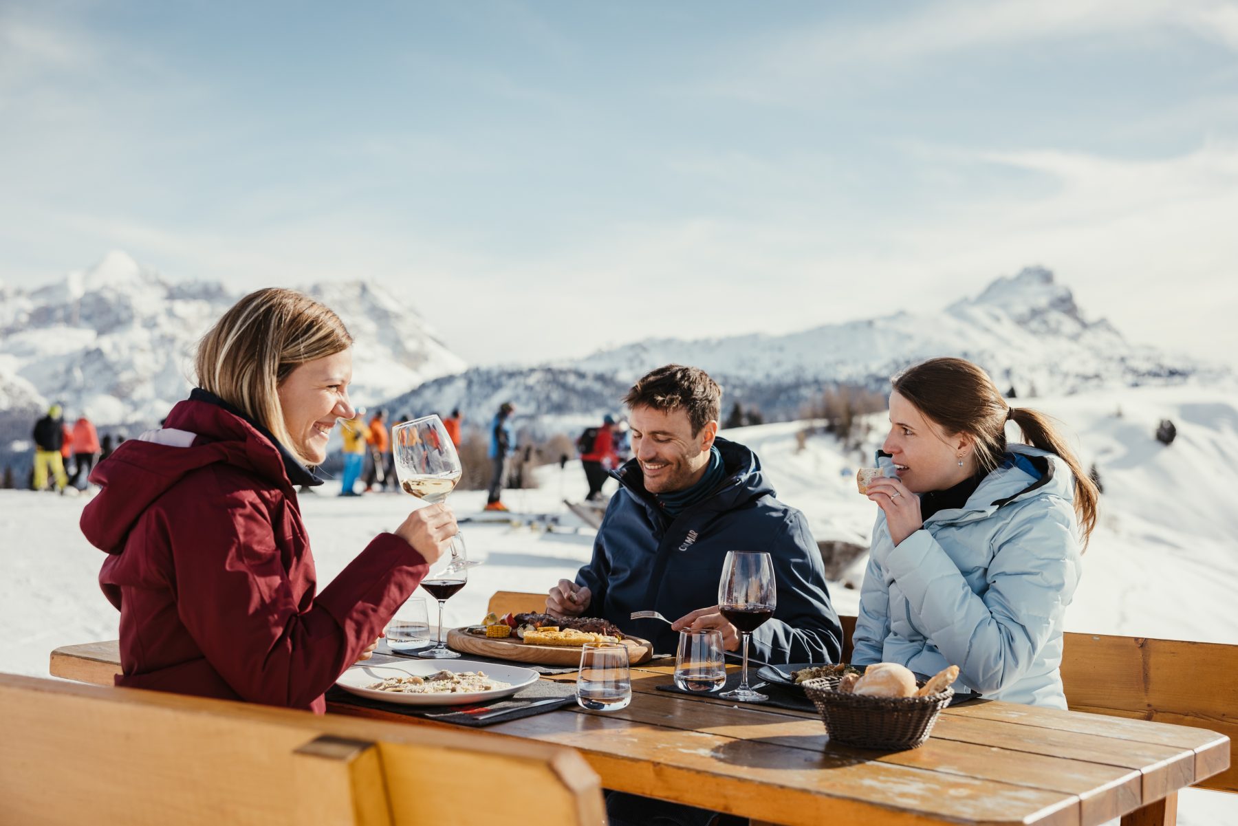 Drie mensen zitten buiten aan tafel te eten en te drinken met op de achtergrond bergen, tijdens de Gourmet Skisafari.