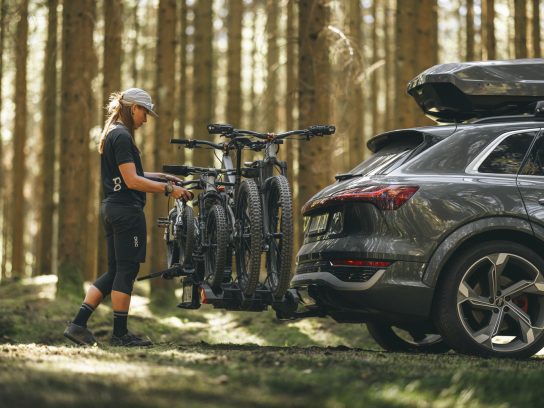 Een vrouw in outdooruitrusting zet mountainbikes vast op de Thule EasyFold 3 fietsendrager achterop een SUV in een bosrijke omgeving.