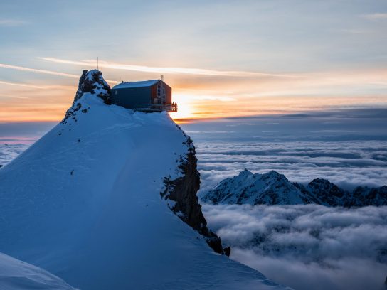 Een berghut bovenop een rotsformatie met uitzicht over een mistig berglandschap met de zonsondergang op de achtergrond.