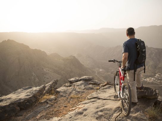 Een man staat naast zijn mountainbike en kijkt over een bergachtig landschap in Oman.