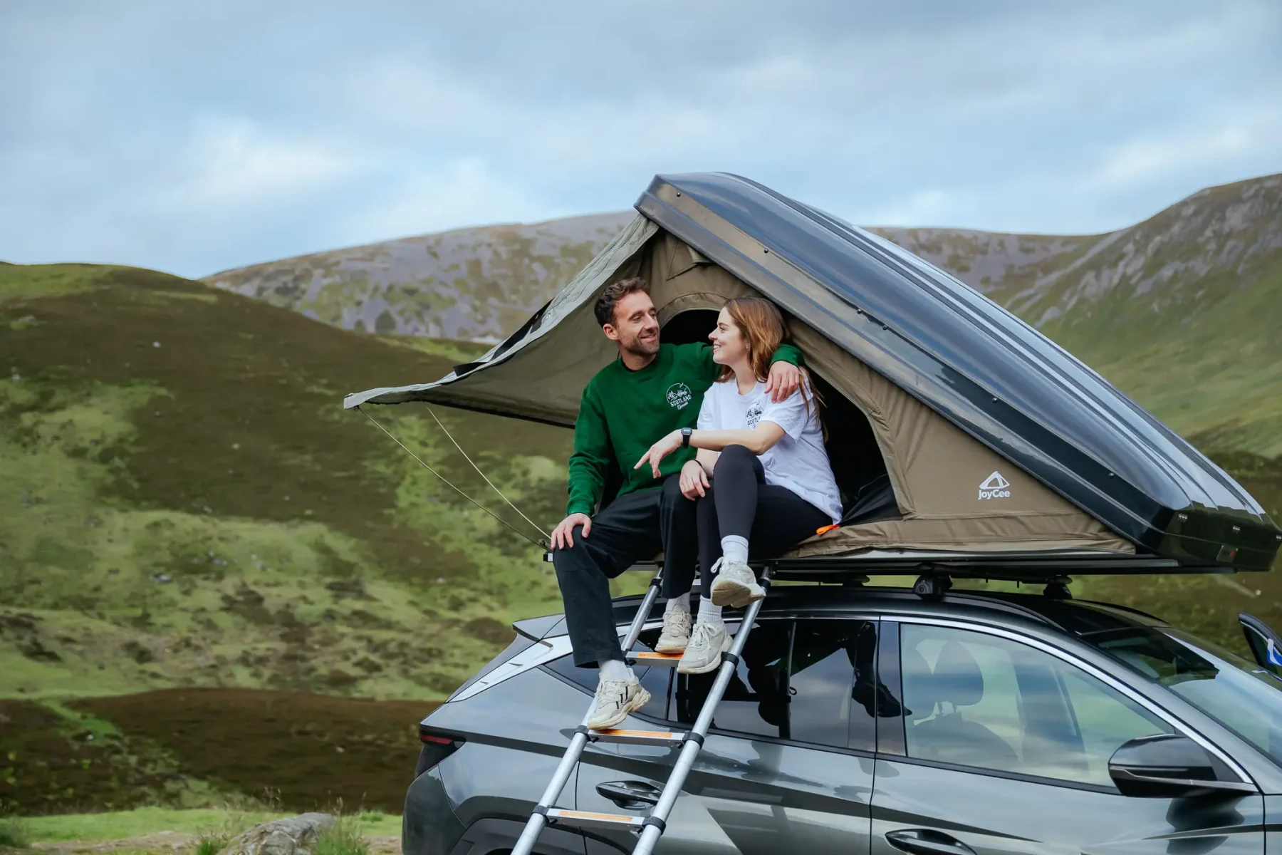 Twee personen in een daktent bovenop een SUV in een heuvelachtig landschap in Schotland tijdens Scotland Nomads.