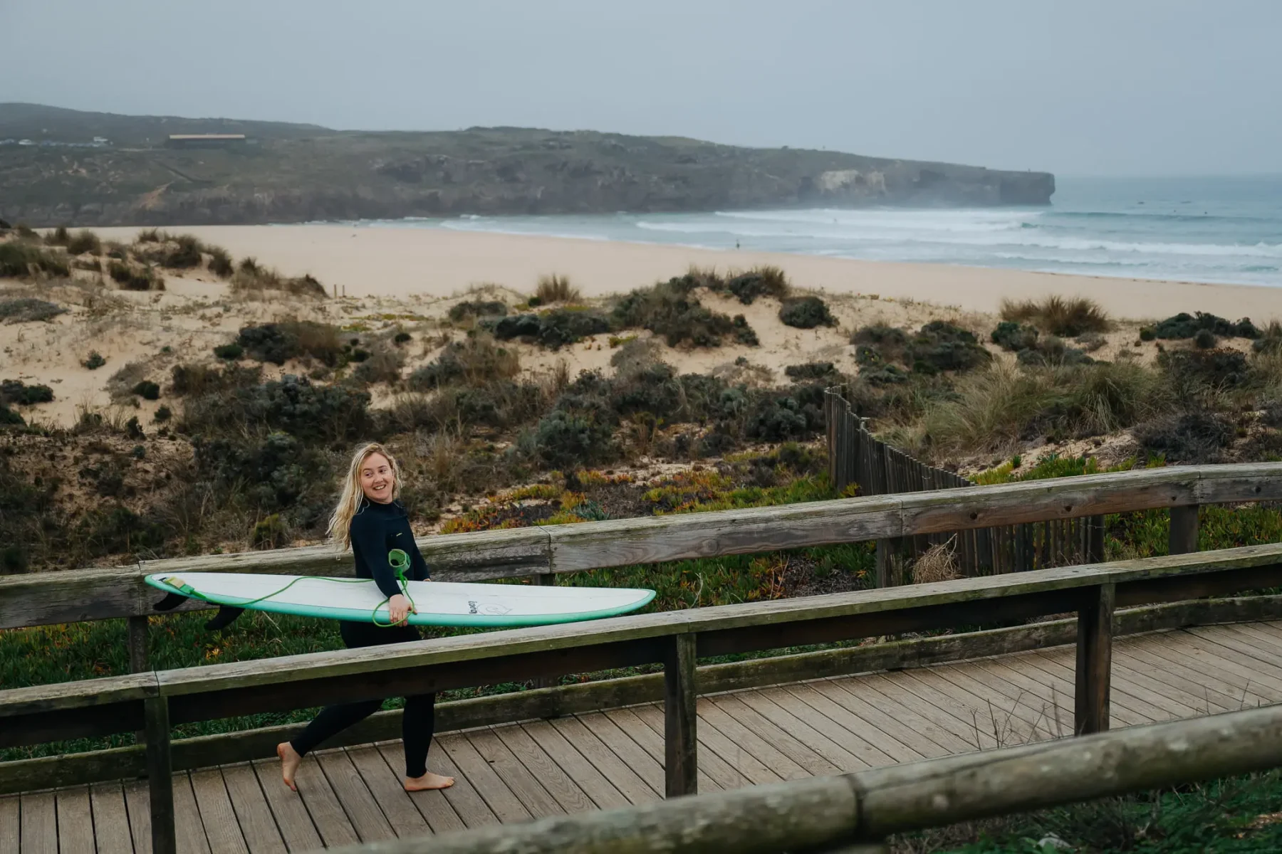 Een vrouw loopt in Portugal over een brug richting de zee met een surfboard onder haar arm.